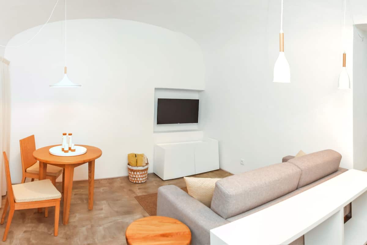 A modern living room with a sofa, a TV mounted on the wall, a wooden dining table with chairs, and a small round table with a bottle.