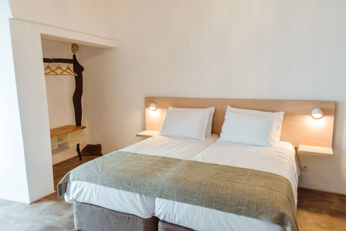 A neatly made bed with a wooden headboard, two bedside tables with wall-mounted lamps, and a hanging garment rack.