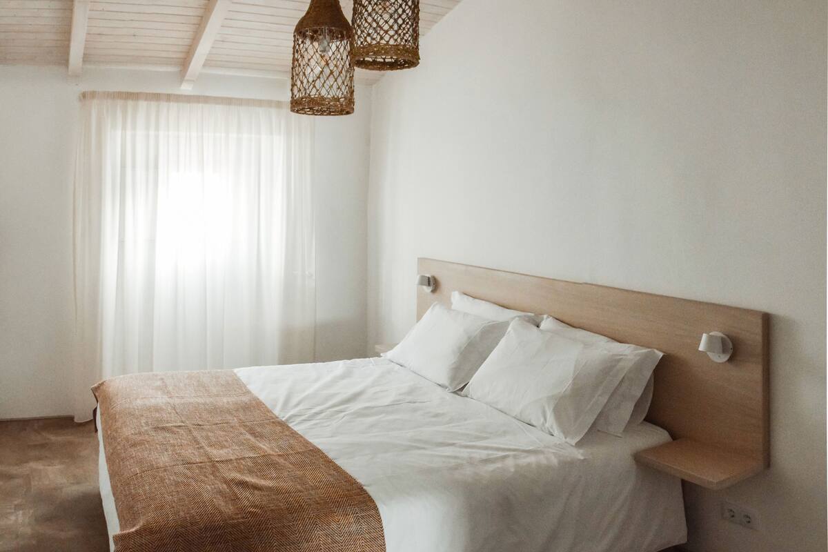 A bedroom with a wooden headboard, a large bed with white linens, two hanging wicker lamps, and a window with sheer curtains.