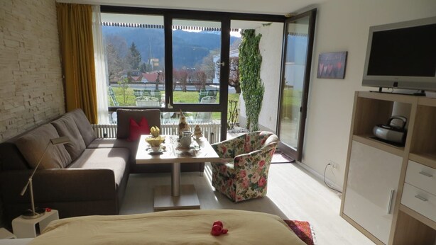 A modern living room with a sofa, a coffee table, a floral armchair, a bed with a floral bedspread, a TV, and a view of a mountainous landscape through large windows. - Apartment - Apartment Kreckel (Schliersee)