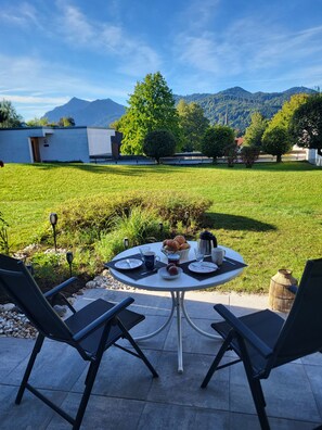 Ein Essbereich im Freien mit einem für zwei Personen gedeckten Tisch, umgeben von Stühlen, mit Blick auf eine Wiese und Berge im Hintergrund.