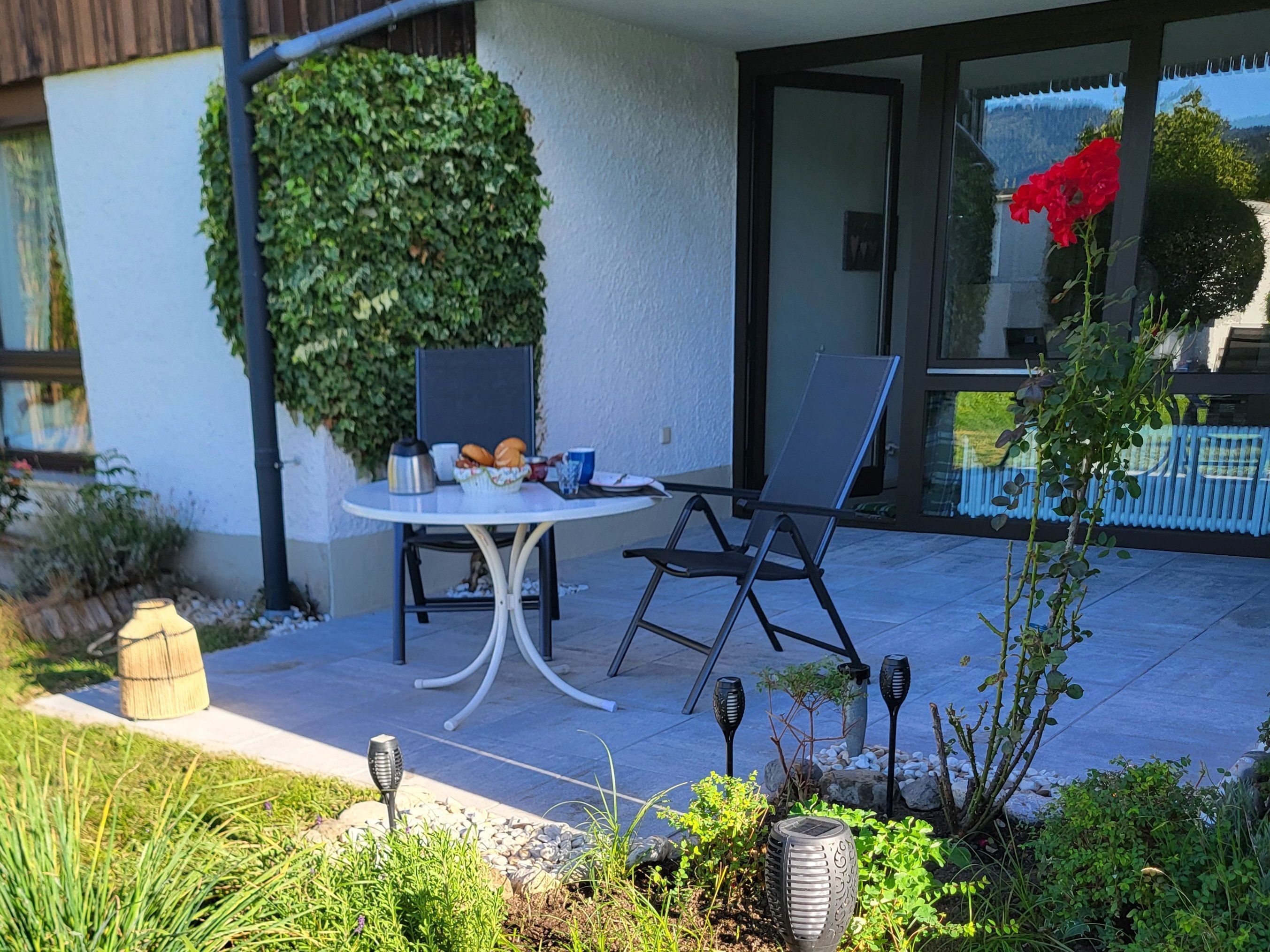 An outdoor patio with a round table, two chairs, a small garden, and a glass door leading to a balcony.