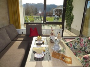 Ein Wohnzimmer mit einer Couch, einem Tisch mit Weihnachtsbaum und Blick auf einen Garten und Berge durch große Fenster.