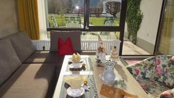 Un salon avec un canapé, une table sur laquelle est placé un sapin de Noël, et une vue sur un jardin et des montagnes, visible à travers de grandes fenêtres.