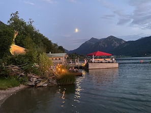 Una estructura de madera junto al lago, con un toldo rojo, rodeada de árboles y montañas.