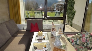 A living room with a sofa, a table with a Christmas tree, and a view of a garden and mountains through large windows. - Apartment - Apartment Kreckel (Schliersee)