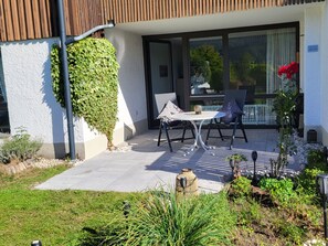 Eine Terrasse mit Tisch und Stühlen, ein kleiner Gartenbereich mit Pflanzen und ein Gebäude mit weißer Fassade und einem Holzbalken-Dach.