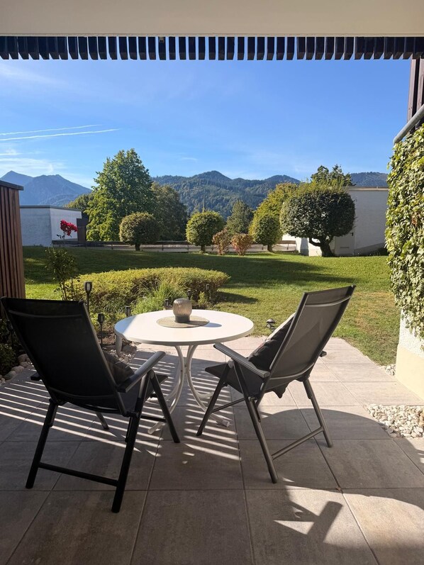 Eine Terrasse im Freien mit zwei Stühlen und einem Tisch, umgeben von einem Garten und Blick auf Berge.