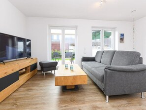 A modern living room with a flat-screen TV, a grey sofa, a wooden coffee table, and a small ottoman.