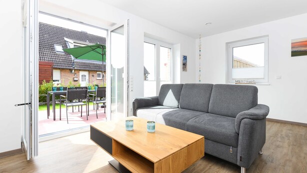 Ein modernes Wohnzimmer mit grauem Sofa, einem Holztisch und einer Glasschiebetür, die auf eine Terrasse im Freien führt.