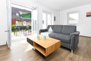 Ein modernes Wohnzimmer mit grauem Sofa, einem Holztisch und einer Glasschiebetür, die auf eine Terrasse im Freien führt.