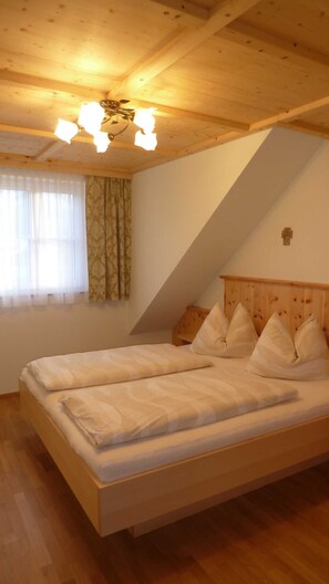 Une chambre avec un plafond en bois, un lit recouvert de draps blancs, une fenêtre ornée de rideaux légers et une croix accrochée au mur.