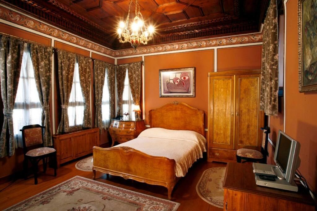 A bedroom with a wooden bed, a desk with a computer, a chair, a wardrobe, and a chandelier.