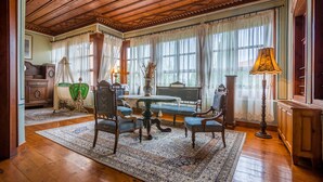 Quarto com teto de madeira, uma grande janela com cortinas leves, uma mesa redonda com cadeiras e um abajur.