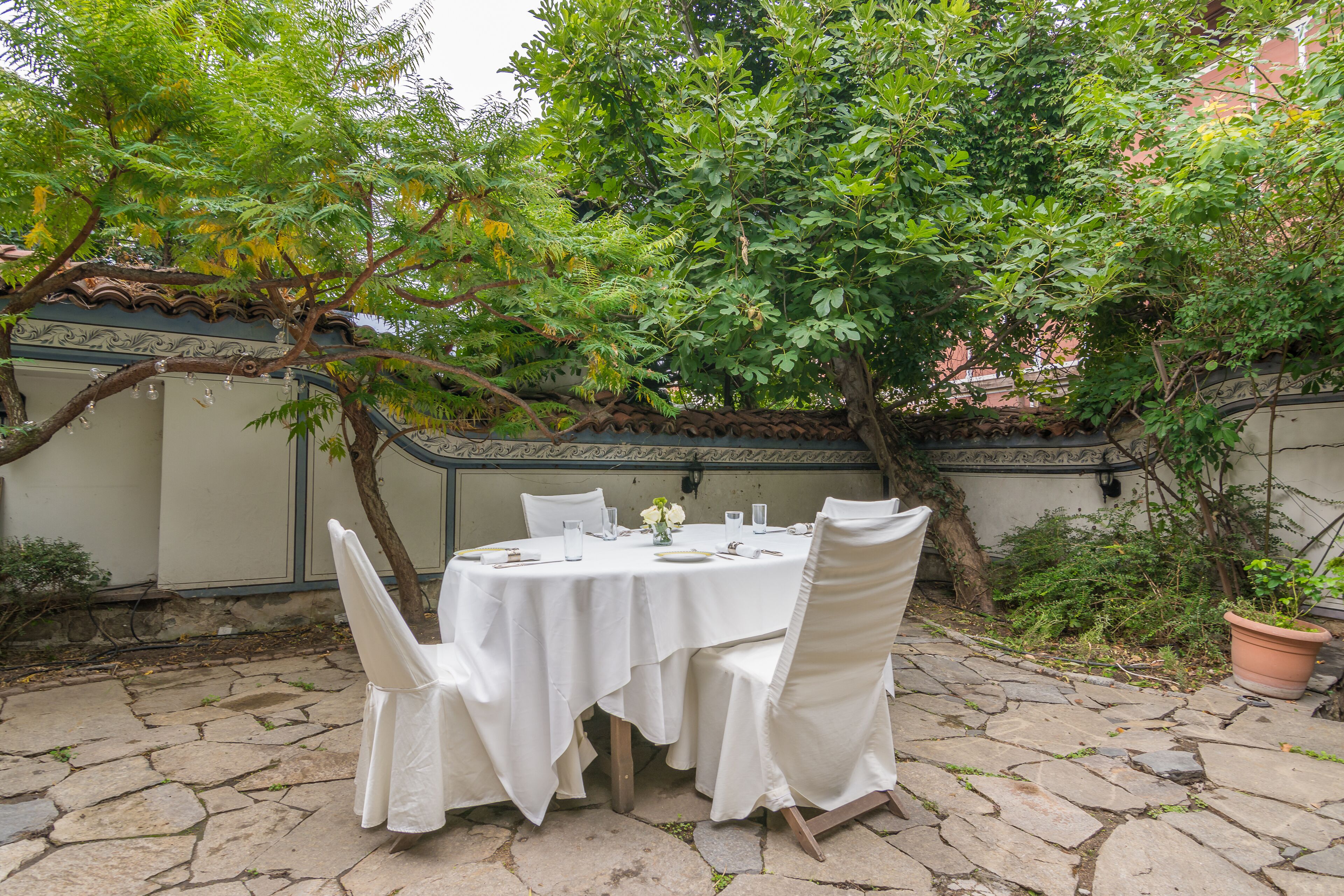 A round table with white tablecloth and chairs, set on a stone patio surrounded by greenery and a building wall.