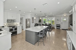 A modern kitchen with white cabinetry, a central island with bar stools, and a dining area with a view of greenery outside.