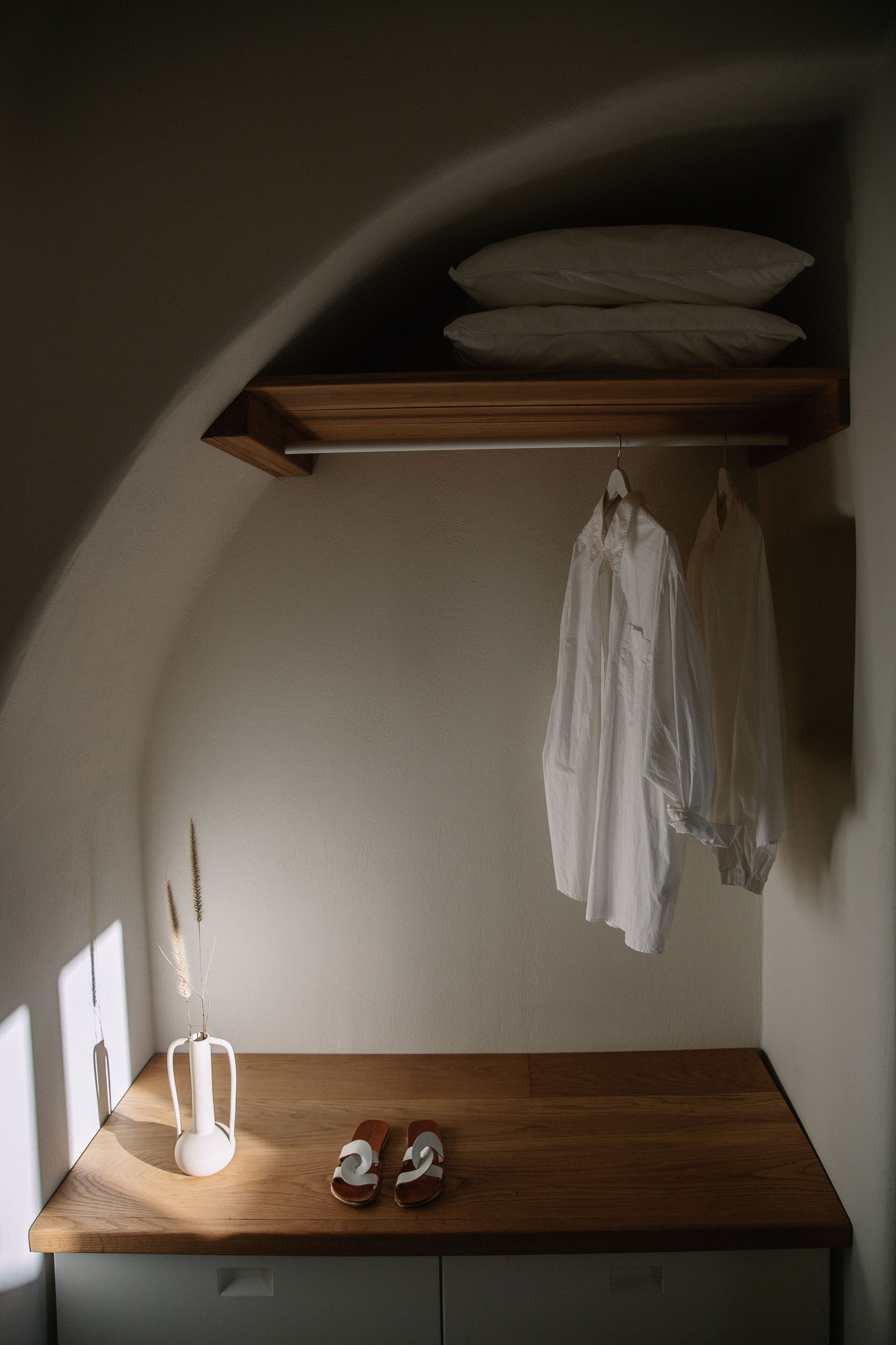A small, dimly lit room with a wooden bench, a white vase with dried plants, and a pair of sandals.