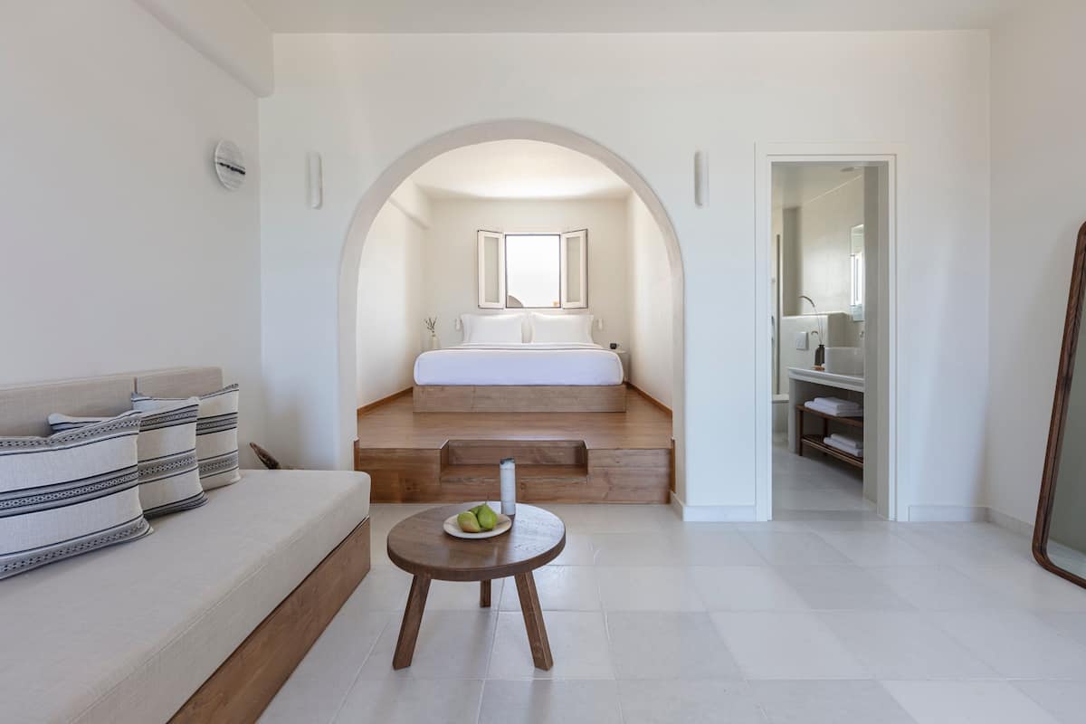 A modern bedroom with a bed, a small table with a plate of fruit, and a bathroom visible through an archway.