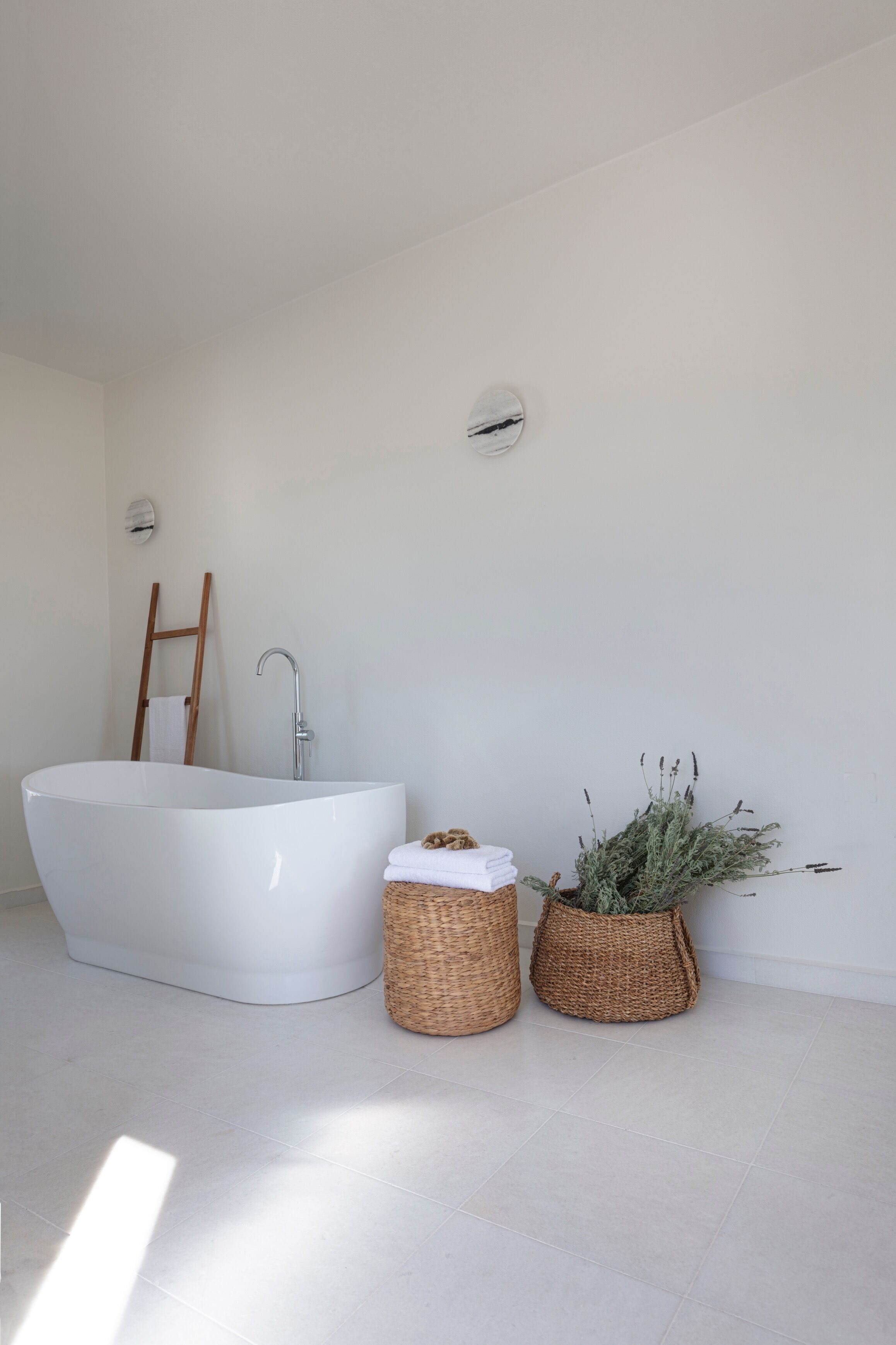 A modern bathroom with a freestanding bathtub, a wooden ladder, and two wicker baskets with towels and plants.