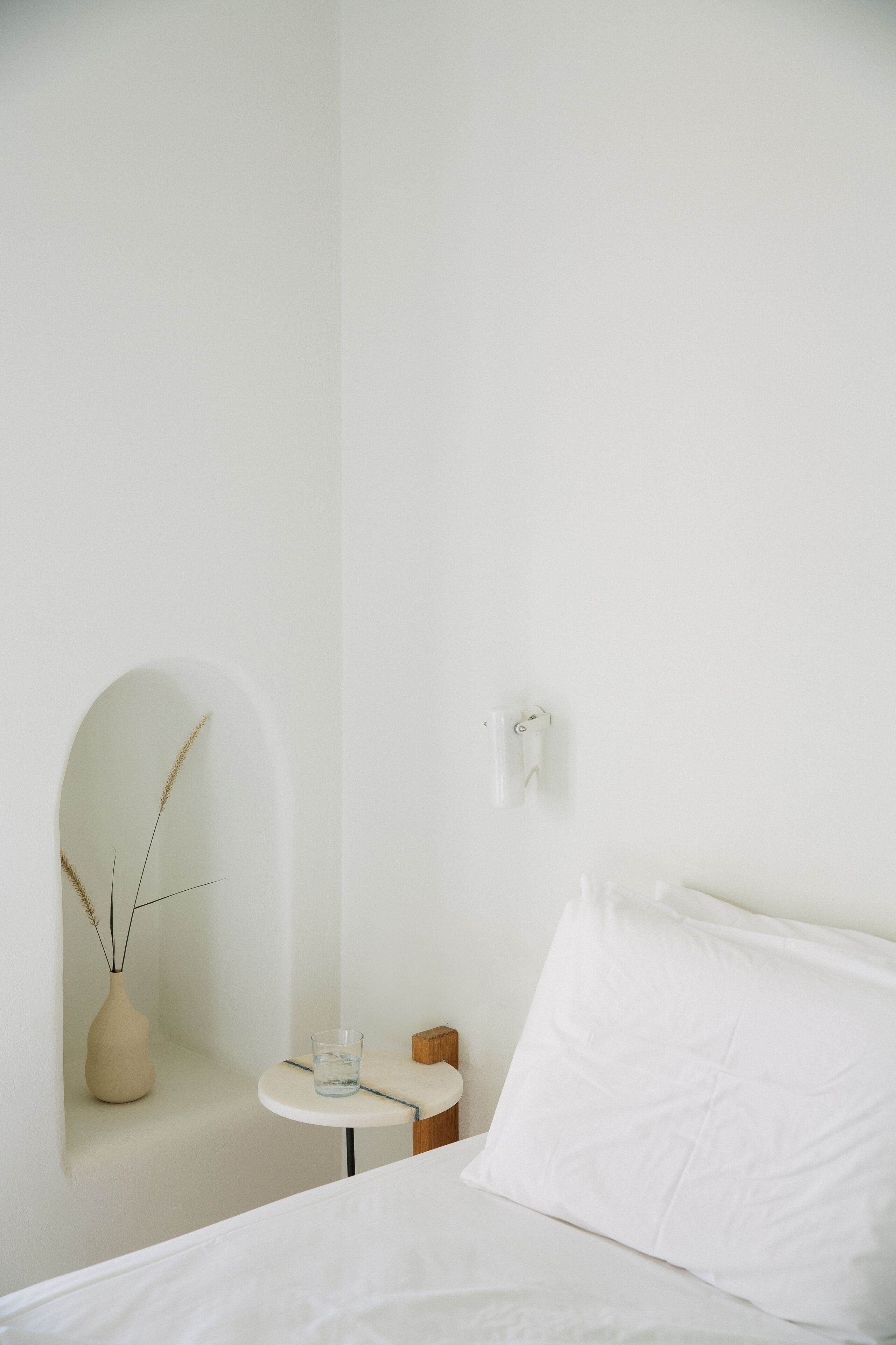 A minimalist room with a bed, a small table with a glass of water, a vase with dried plants, and a wall-mounted light fixture.