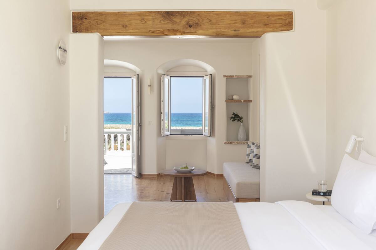 A bedroom with a large bed, a small table with a bowl, a bedside table with a lamp, and a view of the sea through two windows.