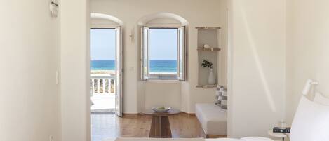 A bedroom with a large bed, a small table with a bowl, a bedside table with a lamp, and a view of the sea through two windows.