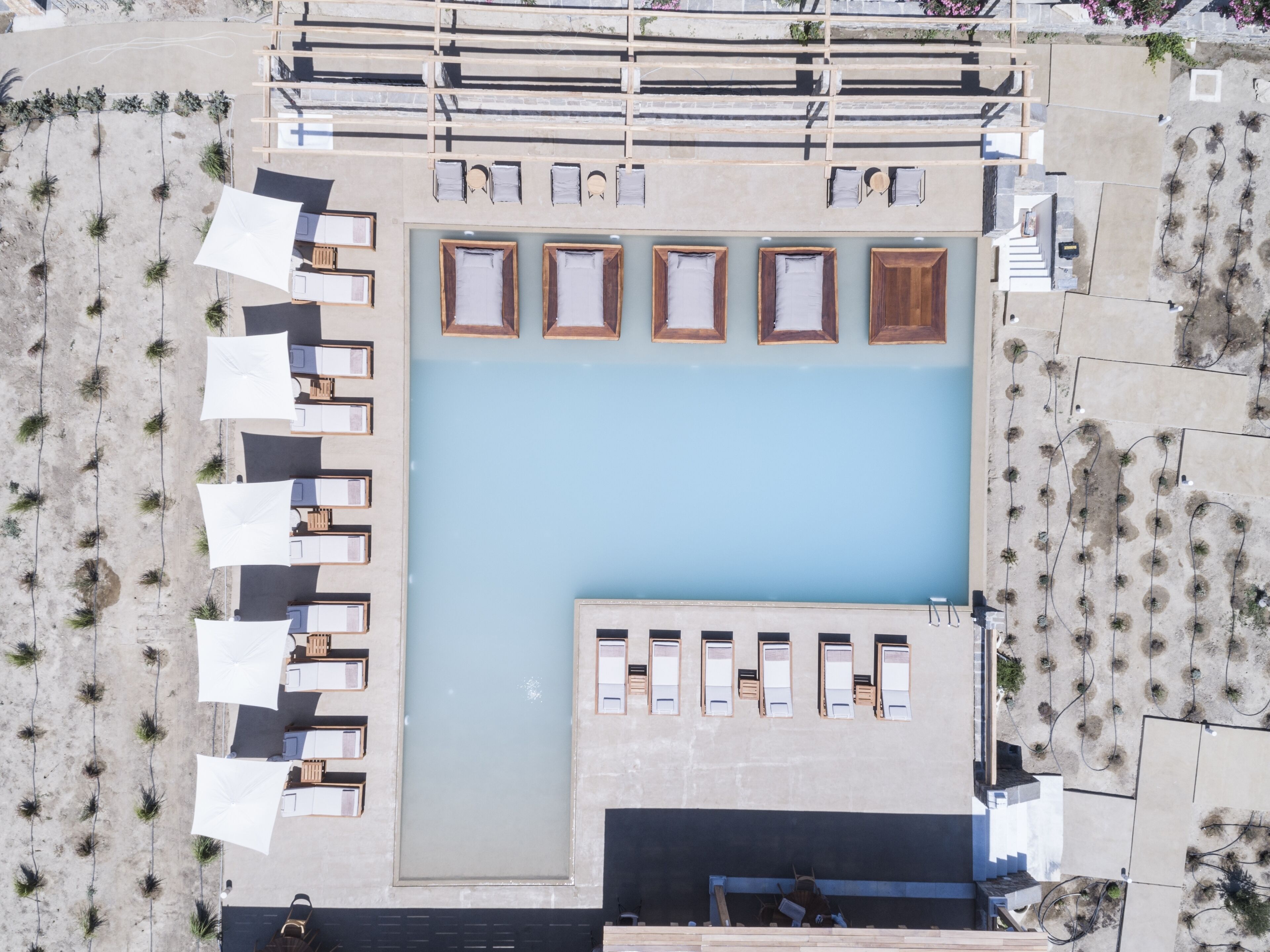 Vista aérea de uma área de piscina com várias espreguiçadeiras e guarda-sóis, rodeada por uma praia de areia e um edifício com fachada azul.