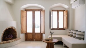 Una habitación con chimenea, un sofá, una mesa redonda y dos ventanas con contraventanas de madera.