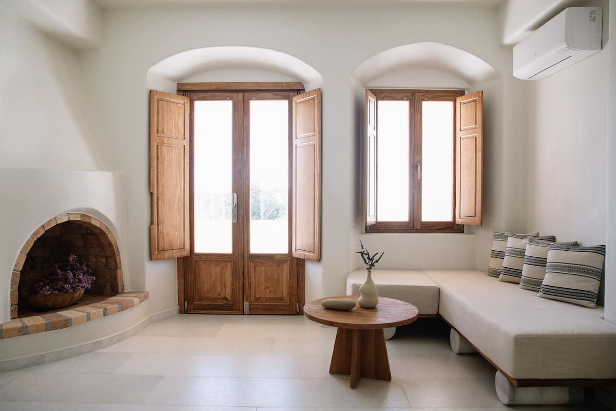 A room with a fireplace, a sofa, a round table, and two windows with wooden shutters.