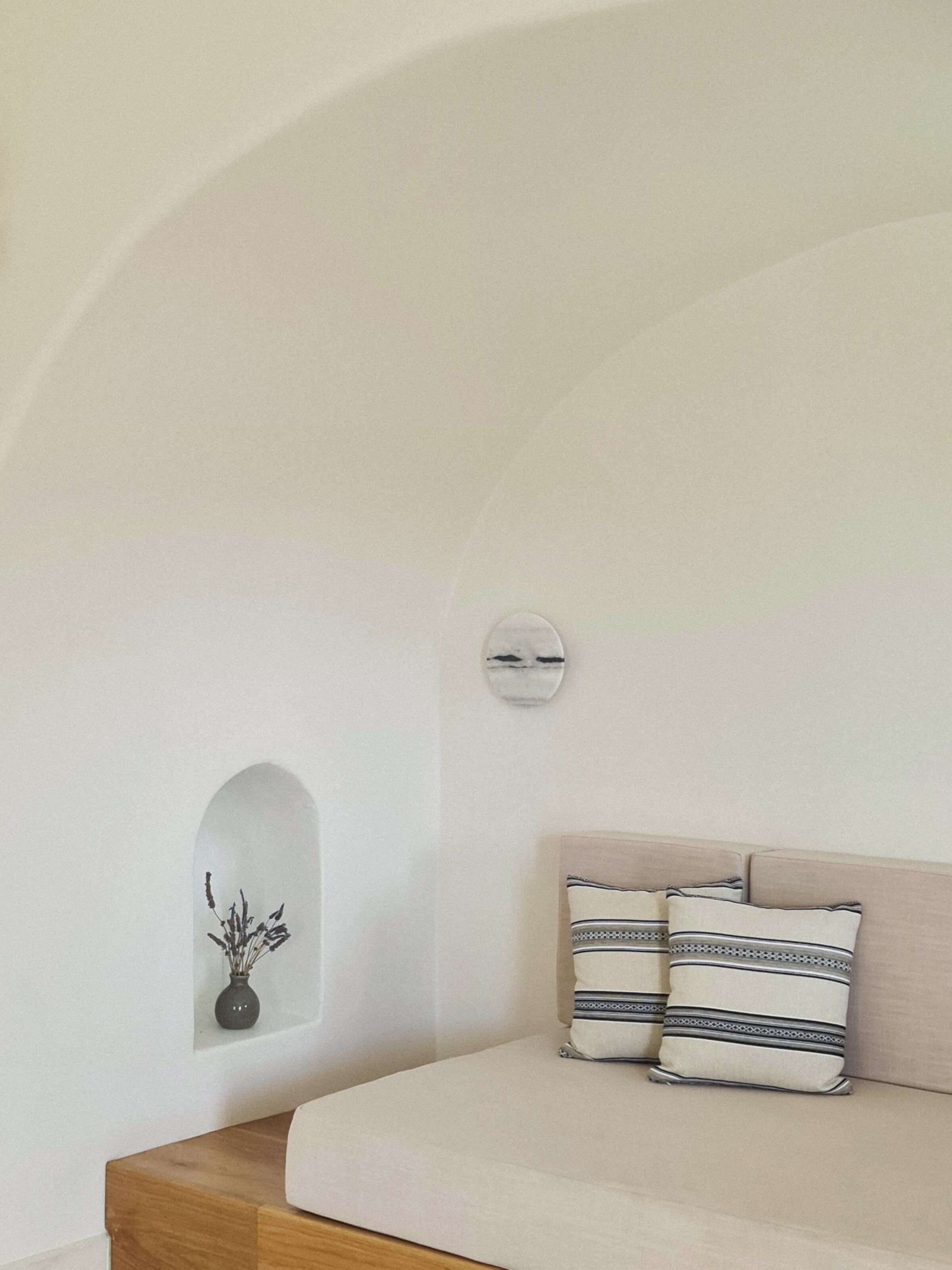 A room with a curved ceiling, a small wall-mounted light, a wooden bedside table, and a beige sofa with patterned pillows.
