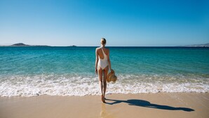Una mujer con traje de baño blanco caminando sobre una playa arenosa con agua azul cristalina y una masa de tierra en la distancia.