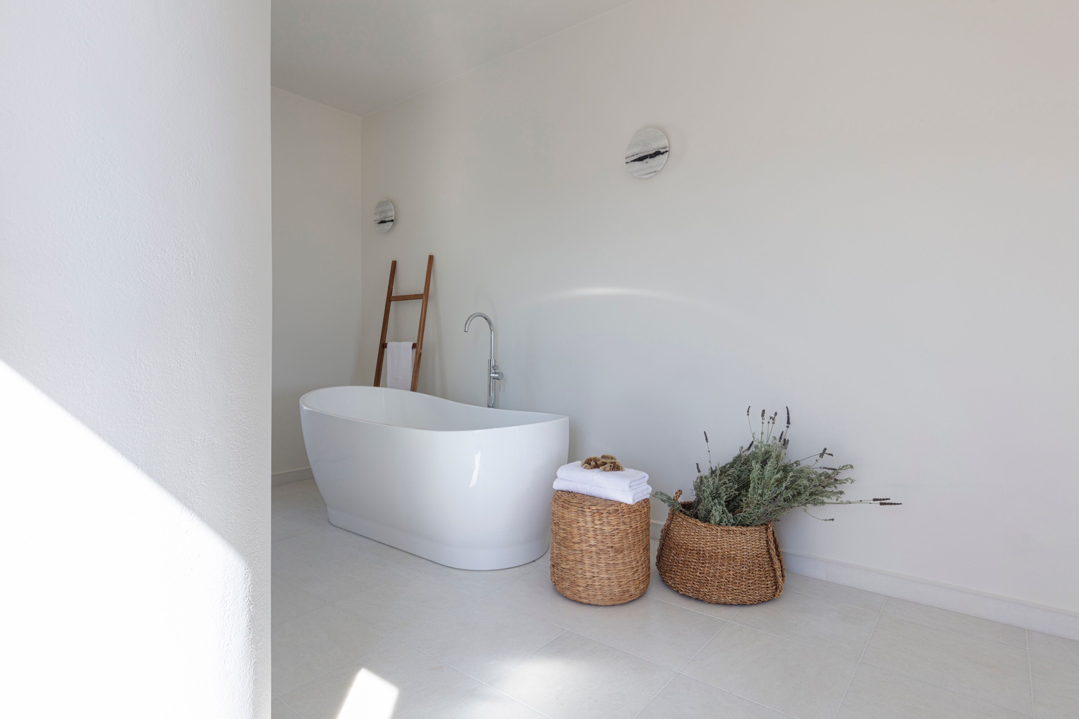 A modern bathroom with a freestanding bathtub, a wooden ladder, and two wicker baskets with plants.