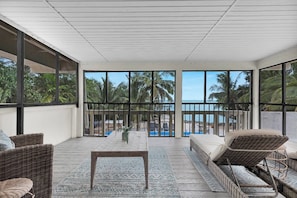 A balcony with wicker furniture, a small table, and a view of the ocean and palm trees.