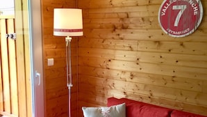 Une pièce chaleureuse avec un canapé rouge, un plaid blanc moelleux et une lampe. On peut également apercevoir un panneau mural en bois et un panneau circulaire.