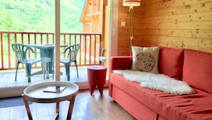 Une pièce chaleureuse avec un canapé rouge, une petite table ronde et deux chaises. Une grande fenêtre offre une vue sur un chalet en bois et des collines verdoyantes.