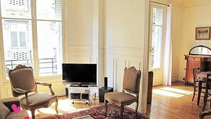 Ein Wohnzimmer mit Flachbildfernseher, zwei Polstersesseln, einem gemusterten Teppich und einem Essbereich mit Holztisch und Stühlen.