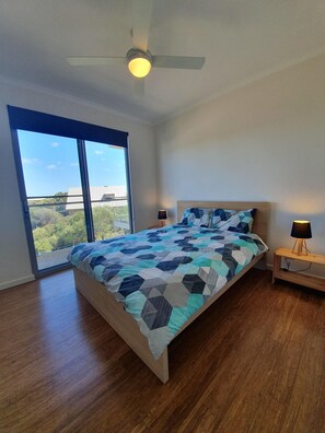A bedroom with a large bed, a ceiling fan, a nightstand with a lamp, and a view through a large window.