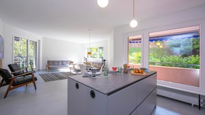 A modern kitchen with a central island, large windows with a view of greenery, and a dining area with a sofa and table. - Coloratura Frontlake Lugano With Terrace (Paradiso)