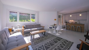 A modern living room with a sofa, coffee table, and a dining area with chairs and a small table. A kitchen with a bar and stools is visible in the background. - Coloratura Frontlake Lugano With Terrace (Paradiso)