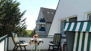 A balcony with two striped chairs, a small table with a flower vase, and a view of a building with a sloped roof. - A 2 GH 2-room balcony (BC.n5) - Stranddomicil Steiner - Guesthouses (Büsum)