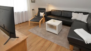 A modern living room with a flat-screen TV, a black leather sofa, a wooden coffee table, and a black armchair with a cushion. - A 2 GH 2-room balcony (BC.n5) - Stranddomicil Steiner - Guesthouses (Büsum)