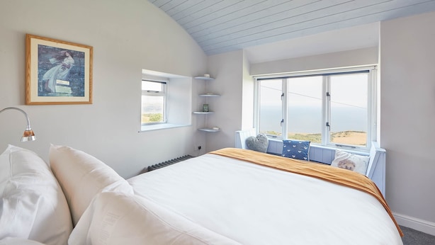 A bedroom with a large bed, a window with a view of the beach, a framed picture on the wall, and a bedside table with a lamp.