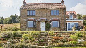 A stone house with a tiled roof, a front door, and a small garden with steps leading to the entrance.