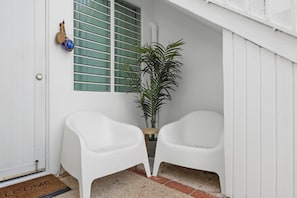 Una puerta blanca con un felpudo que dice 'WELCOME', una planta en maceta y dos sillas de plástico blancas.