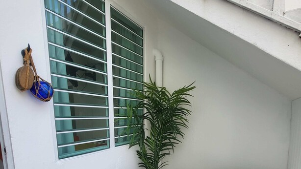 Un petit espace extérieur clos, avec un mur blanc, une fenêtre dotée de stores horizontaux, une plante en pot et deux chaises en plastique blanches.
