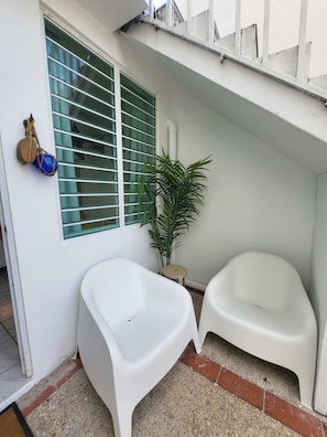 A small, enclosed outdoor area with a white wall, a window with horizontal blinds, a potted plant, and two white plastic chairs.