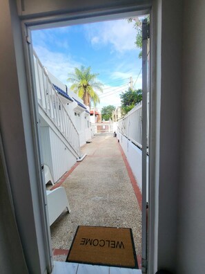 Un sendero estrecho que lleva a un edificio con paredes blancas y techo azul, un felpudo en la entrada y una palmera al fondo.