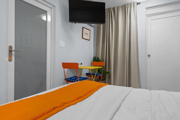 A bedroom with a bed, a TV mounted on the wall, a small table with chairs, and a potted plant.