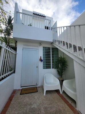 A two-story building with a white exterior, a balcony, a white door, a small white chair, a welcome mat, and a staircase with decorative tiles.