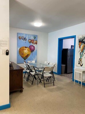 Un comedor con una mesa de vidrio rodeada de sillas, un cuadro de globos en la pared y una puerta que da a otra habitación.